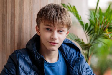 Açık verandadaki ahşap bir sütuna yaslanmış mavi kabarık ceketli, ciddi bir gencin yakın plan portresi.