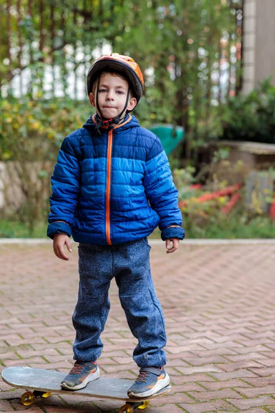 Sıcak mavi ceketli ve kasklı genç bir çocuk sonbaharda tuğladan bir kaldırımda kaykay sürmeyi öğreniyor.