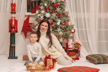 Kırmızı ve beyaz süslemeler ve hediyelerle süslenmiş bir Noel ağacının yanında rahat krem süveter içinde oturan mutlu anne ve küçük oğlu.