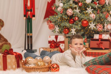 Mutlu genç bir çocuk beyaz kabarık bir halıya uzanmış, Noel süslerinin yanında poz veriyor ve parlak süslenmiş bir ağacın önünde hediyeler veriyor.