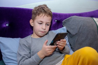 Odaklanmış genç bir çocuk yatakta rahat bir şekilde uzanıyor, oyun ya da eğlence için akıllı telefon kullanıyor parlak mor bir yatak başlığına karşı.