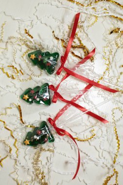 Üç noel ağacı şekilli lolipoplar ya da kırmızı kurdelelerle bağlanmış şekerlemeler, beyaz ve altın renginde konfetiler üzerinde duruyor.