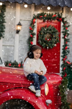 Kırmızı bir kamyonun kaputunda oturan tüylü beyaz kazak ve kulaklıklı mutlu genç kız hafif bir kar yağışı altında gülümsüyor.
