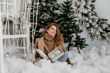 Deve kürkü ve örgü kazak giyen mutlu genç bir kadın yapay karda oturuyor, neşeli bir gülümsemeyle beyaz bir noel hediyesi kutusu açıyor.