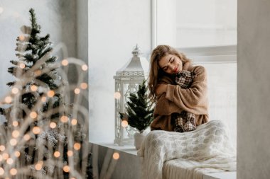 Sakin, genç bir kadın, bir yastığa sarılmış ve bir battaniyeye sarılmış, güneşli bir pencere pervazında oturuyor.