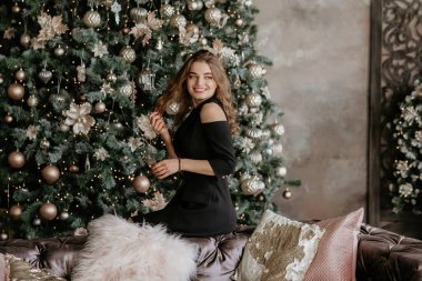 Siyah bir gece elbisesi içinde neşeli genç bir kadın kadife bir koltukta oturur ve güzelce süslenmiş bir Noel ağacına bakar.