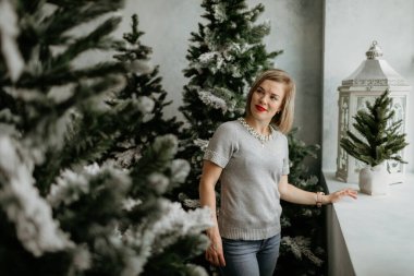 Gri kazaklı ve inci kolyeli şık bir kadın gülümsüyor, kar tozlu Noel ağaçları ve beyaz bir fenerle pencerenin kenarında duruyor..