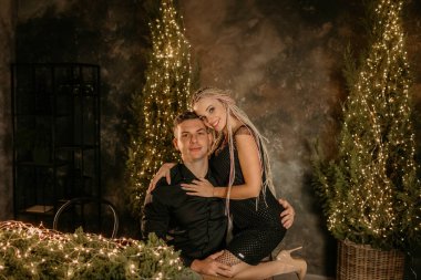 Mutlu ve şık bir çift Noel çelenkleri ve ışıklarla süslenmiş bir masada otururken kameraya bakarken sarılıyor.