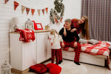 Kırmızı ekose pijamalı mutlu bir aile beyaz bir dolabın yanında poz veriyor ve genç kız Noel çorabında hediye arıyor.