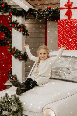 Beyaz örgü elbiseli neşeli bir kız beyaz bir arabanın kaputunda mutlu bir şekilde oturuyor, dekoratif karda ellerini kaldırıyor.