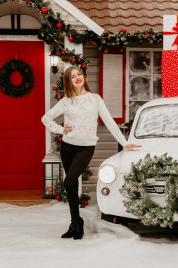 Beyaz örgü süveter ve siyah çizmeler giyen neşeli bir kadın, karlı bir Noel ortamında klasik beyaz bir arabanın yanında güven içinde poz veriyor.