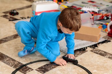 Parlak mavi eşofmanlı genç bir çocuk parlak fayanslı zeminde diz çöker, iyi aydınlatılmış bir odada oyuncak tren raylarını dikkatle bağlar.