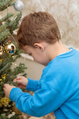 Mavi gömlekli genç bir çocuğun dikey yakın çekimi. Kapalı bir Noel ağacının dalına altın bir süs asmaya odaklanmış.