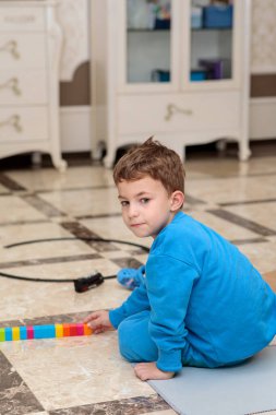 Mavi eşofmanlı, parlak fayanslı bir zeminde oturan ve doğrudan kameraya bakan sevimli bir çocuğun dikey portresi.