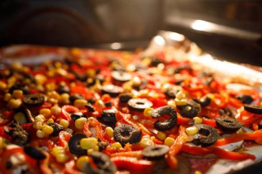 Renkli sebzelerle, siyah zeytinlerle ve mısırla kaplı ev yapımı bir pizzanın aşırı yakın plan çekimi. Parlak bir fırının içinde pişiyor.