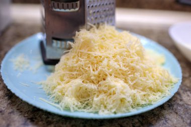 Açık mavi tabakta, granit mutfak tezgahına kurulmuş taze rendelenmiş sarı peynir yığınının üzerinde duran paslanmaz çelik kutunun yakın görüntüsü.