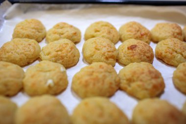 Yukardan gelen sıcak ve yoğun ışıkla aydınlatılan, parşömen kağıdının üzerinde duran altın, yeni pişmiş peynirli pufların yakın çekimi.