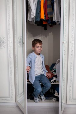 Mavi çizgili gömlek ve kot pantolonlu genç adam açık beyaz bir gardıropta oturuyor, dağınık bir alanda kameradan dikkatle başka yere bakıyor.