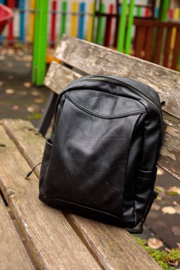 Yıpranmış ahşap bir park bankında fermuarlı modern siyah deri sırt çantası seyahat, eğitim, öğrenci yaşam tarzı ya da her gün dışarı çıkma kavramı.