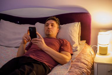 Yetişkin bir adam yatakta yatar ve uyumadan önce akıllı telefon kullanır, erkek sosyal medyada kayar ya da rahat bir yatak odasında gece lambasıyla mesajlaşır.
