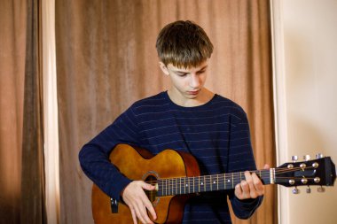 Koyu renk çizgili süveter giyen genç müzisyen evde akustik gitar çalıyor müzik becerileri, yaratıcı hobi ve eğitim kavramı üzerinde çalışan dalgın ergen.