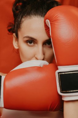 Kırmızı boks eldivenli genç bir kadının yoğun portresi yüzünü örtüyor ve kameraya kararlılıkla bakıyor. Stüdyoda kırmızı arka planlı spor eğitimi alıyor.