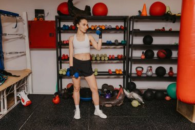 Spor giyimli, formda genç bir kadın spor salonunda mavi halterlerle duruyor, bayan sporcu ağırlık, spor salonu ve sağlık konseptiyle antrenman için hazırlanıyor.