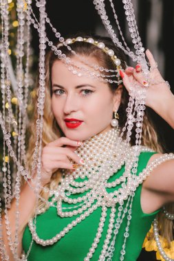 Kırmızı dudaklı, kristal boncuk süslemelerden bakan, inci kolyeli, yeşil elbiseli, zarif tatil güzeli ve moda konsepti olan çekici bir kadının portresini yakından çek.