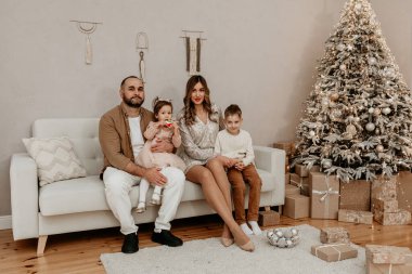 Dört kişilik mutlu bir aile, süslü bir Noel ağacının yanında beyaz bir kanepede oturuyor. İki çocuklu ebeveynler, kış tatilini sıcak bir evde birlikte kutluyorlar.