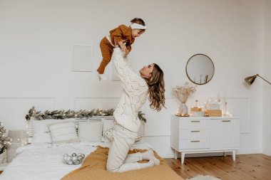 Mutlu anne bir yatakta diz çöküp kızını havaya kaldırıyor. Kış tatillerinde parlak bir yatak odasında eğleniyor ve oyun oynuyor. Annelik kavramının neşesi.