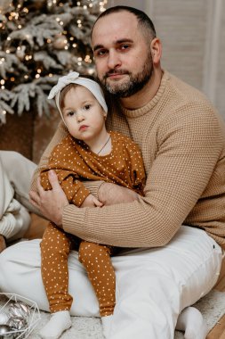 Sevgi dolu bir baba yerde oturup, Noel ağacının yanında küçük kızına nazikçe sarılıyor. Sıcak aile bağı ve kış tatillerinde babalık kavramı.