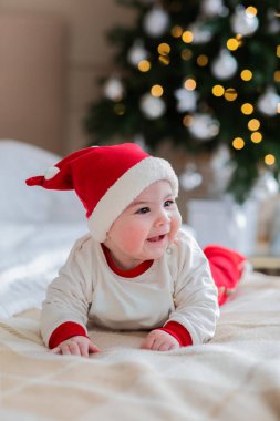 Kırmızı Noel Baba şapkalı sevimli bebek, karnının üstünde yatıyor ve süslenmiş bir Noel ağacının yanında gülümsüyor. Işıkları olan neşeli bebek evde kış tatilinin tadını çıkarıyor.