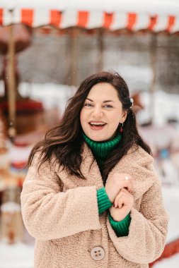 Kahkaha atan bej ceketli ve yeşil balıkçı yakalı bir kadının portresi. Kış atlıkarıncaya yakın, neşeli bir duygu ve karlı bir parkta bayram havası.