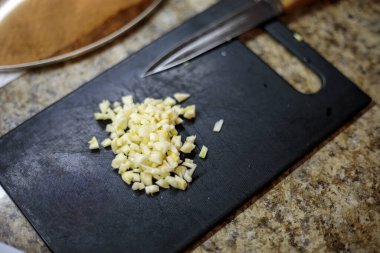 Arka planda şef bıçağı olan ve ev yapımı yemek için aromatik malzemeler hazırlayan siyah kesme tahtasının üzerinde ince doğranmış taze sarımsağı kapat.
