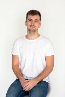 Beyaz t-shirt ve kot giyen yakışıklı genç adam beyaz arka planda stüdyo resminde sandalyede oturuyor.