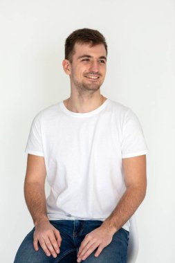 Beyaz t-shirt ve kot giyen yakışıklı genç adam beyaz arka planda stüdyo resminde sandalyede oturuyor.