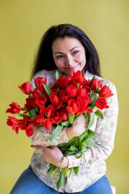 Uzun siyah saçlı güzel bir kadın büyük bir buket kırmızı lale tutuyor ve bahar tatili için stüdyo resminde sarı arka planda gülümsüyor. 8 Mart, Kadınlar Günü, Anneler Günü, Doğum günü