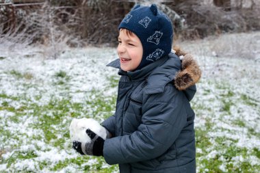 Mutlu çocuk kışın başında dışarıda kartopu tutuyor, çocuk şehir bahçesinde ilk karla oynuyor, çocukluk kış eğlencesi, mevsimsel açık hava aktivitesi, soğuk hava yaşam tarzı