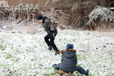 Karlı arka bahçede oynayan iki kardeş, küçük çocukla ahşap kızak çeken büyük çocuk, kış eğlencesi, aile eğlencesi, açık havada çocukluk eğlencesi.