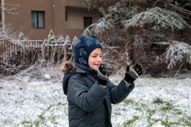 Kışın açık havada ilk kar yağışının keyfini çıkaran çocuk, kürk kapüşonlu ve eldivenli kalın bir ceket giyiyor, parkta gülümsüyor ve kar tanelerini yakalıyor, soğuk havada doğal çocukluk anı.