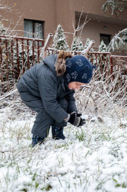 Kışın açık havada ilk kar yağışının keyfini çıkaran çocuk, kürk kapüşonlu ve eldivenli kalın bir ceket giyiyor, parkta gülümsüyor ve kar tanelerini yakalıyor, soğuk havada doğal çocukluk anı.
