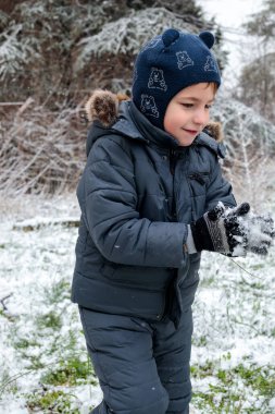 Kışın açık havada ilk kar yağışının keyfini çıkaran çocuk, kürk kapüşonlu ve eldivenli kalın bir ceket giyiyor, parkta gülümsüyor ve kar tanelerini yakalıyor, soğuk havada doğal çocukluk anı.