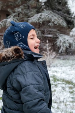 Kışın açık havada ilk kar yağışının keyfini çıkaran çocuk, kürk kapüşonlu ve eldivenli kalın bir ceket giyiyor, parkta gülümsüyor ve kar tanelerini yakalıyor, soğuk havada doğal çocukluk anı.
