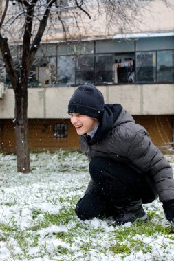 Karlı çimenlerde çömelmiş ve kış parkında yüksek sesle gülen mutlu genç çocuk. Neşeli genç soğuk havada dışarıda karla eğleniyor..