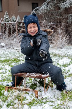 Küçük mutlu çocuk ahşap bir kızağın üzerinde oturuyor ve dışarıya kartopu atıyor. Sıcak kış kıyafetleri içinde parkta karla oynayan tatlı bir çocuk..