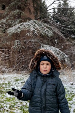 Kışın açık havada ilk kar yağışının keyfini çıkaran çocuk, kürk kapüşonlu ve eldivenli kalın bir ceket giyiyor, parkta gülümsüyor ve kar tanelerini yakalıyor, soğuk havada doğal çocukluk anı.