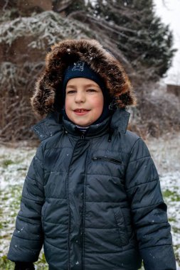 Kışın açık havada ilk kar yağışının keyfini çıkaran çocuk, kürk kapüşonlu ve eldivenli kalın bir ceket giyiyor, parkta gülümsüyor ve kar tanelerini yakalıyor, soğuk havada doğal çocukluk anı.
