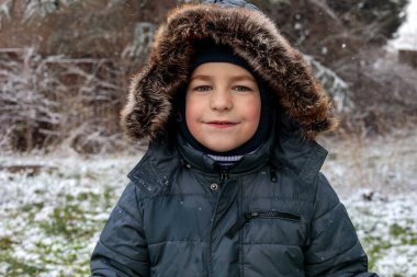 Kışın açık havada ilk kar yağışının keyfini çıkaran çocuk, kürk kapüşonlu ve eldivenli kalın bir ceket giyiyor, parkta gülümsüyor ve kar tanelerini yakalıyor, soğuk havada doğal çocukluk anı.