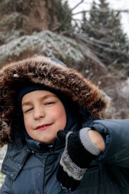 Kışın açık havada ilk kar yağışının keyfini çıkaran çocuk, kürk kapüşonlu ve eldivenli kalın bir ceket giyiyor, parkta gülümsüyor ve kar tanelerini yakalıyor, soğuk havada doğal çocukluk anı.