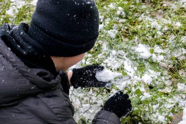 Genç çocuk kışın dışarıda ilk karla oynuyor. Sıcak ceket giyen ergen, şapkalı ve eldivenli genç yeşil çimlerde taze kara dokunuyor. Soğuk havada sezonluk gençlik anıları.